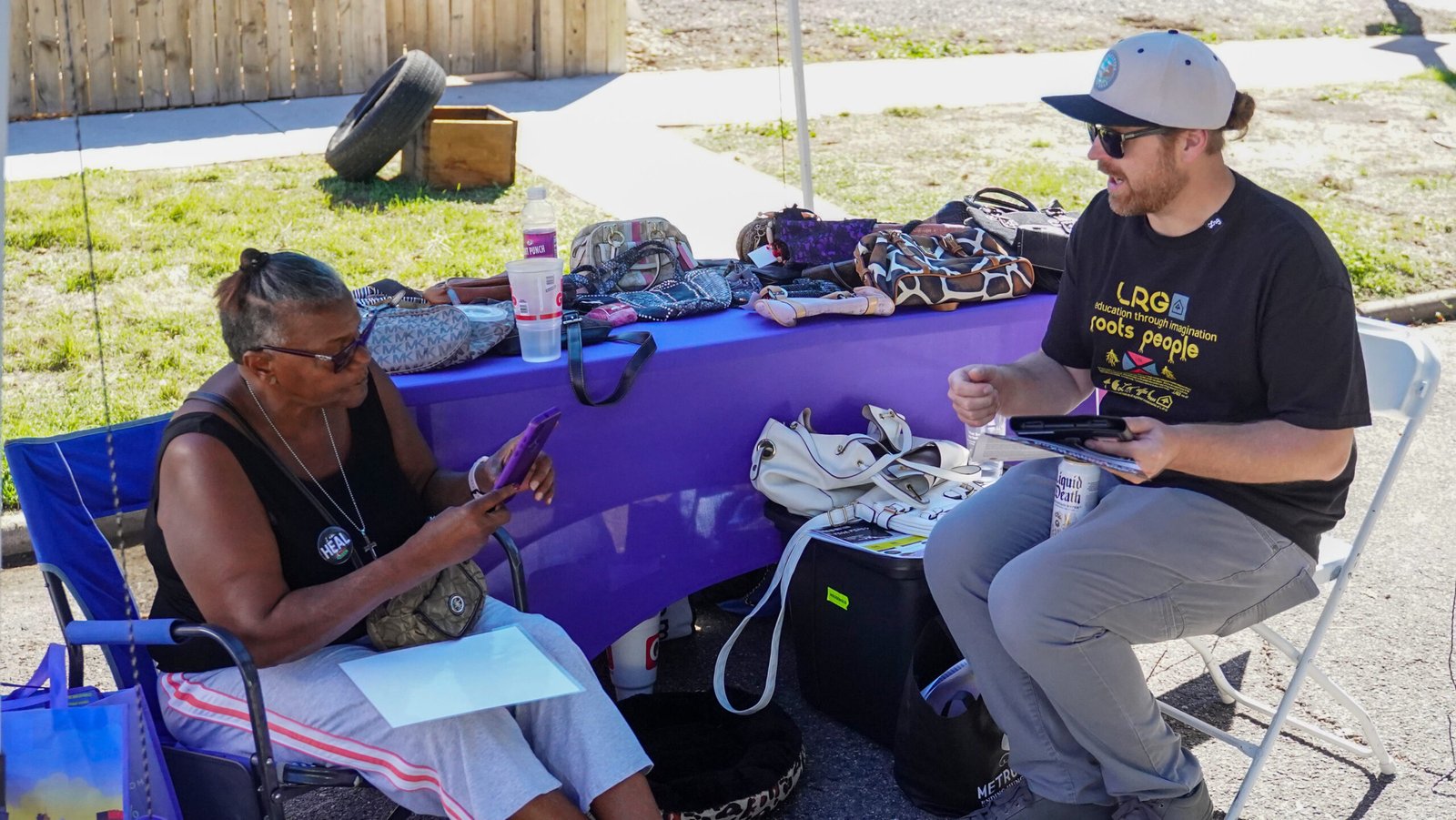 hs10 Shan and a community member sit in lawn chairs in front of a vendor's table.