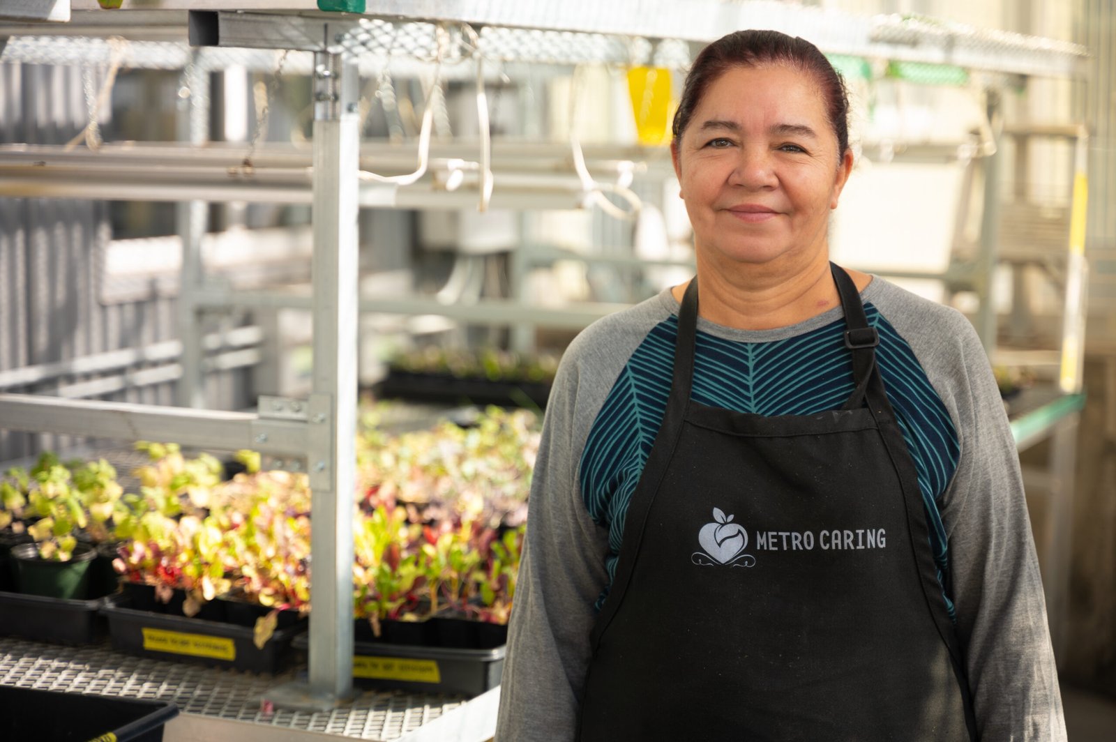 Blanca smiles standing in the Metro Caring greenhouse and wearing a black Metro Caring apron.