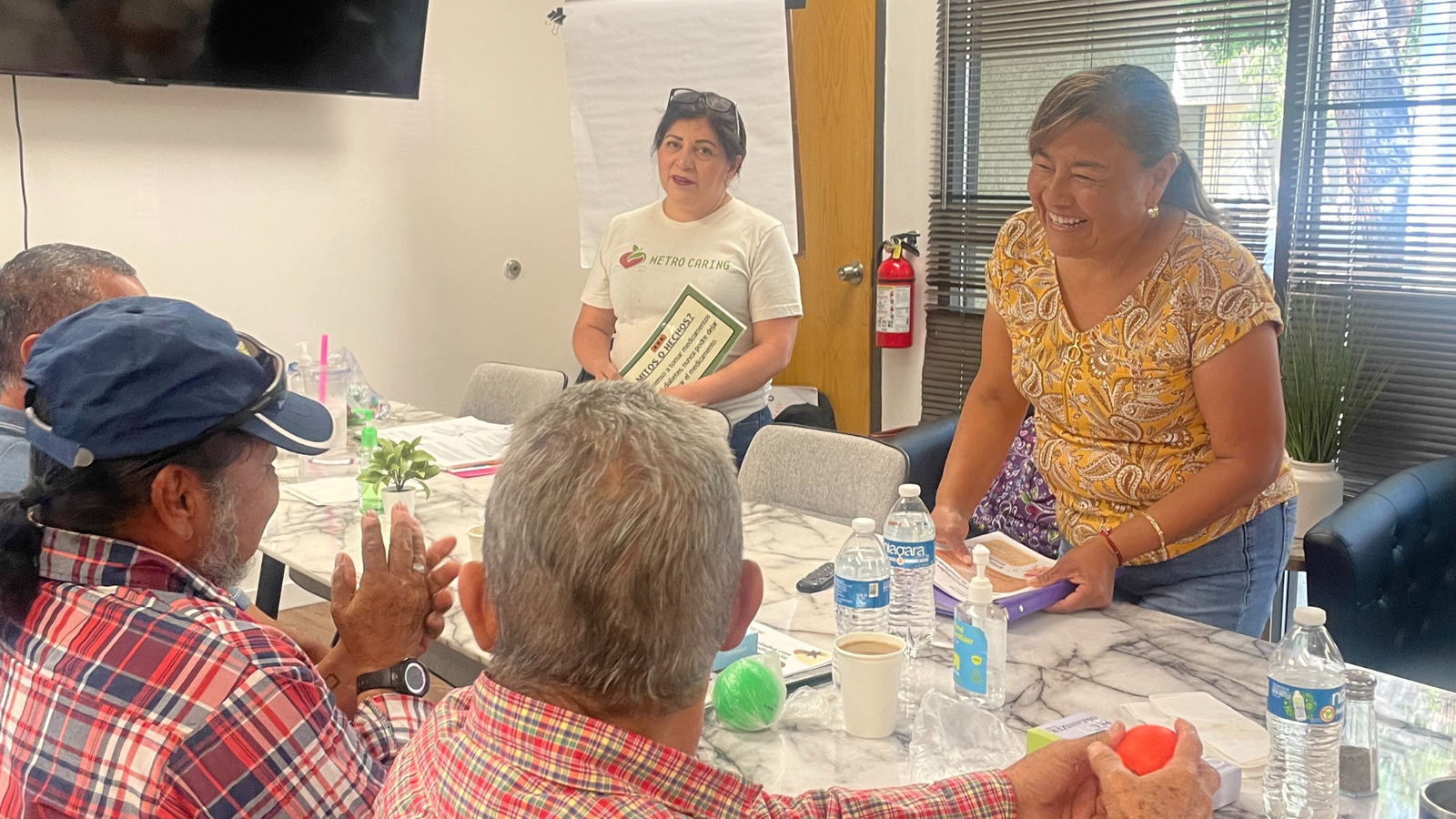 Latina woman holds a packet of papers, smiling, while she stands at a table talking to several people in Metro Caring's Diabetes Among Friends support group/